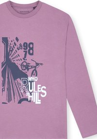 Chemise à manches longues lavande avec un design graphique représentant un vélo et le texte "QUI RÈGNE SUR LA COLLINE" en bleu foncé contrastant.