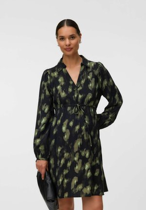 Donna con capelli scuri raccolti in un chignon, che indossa un abito a maniche lunghe nero e verde con fantasia, che tiene una pochette nera su uno sfondo neutro.