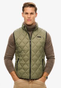 Groene gewatteerde vest met een hoge kraag en ritssluiting, voorzien van zijzakken en zwarte pipings, gedragen over een bruine coltrui.