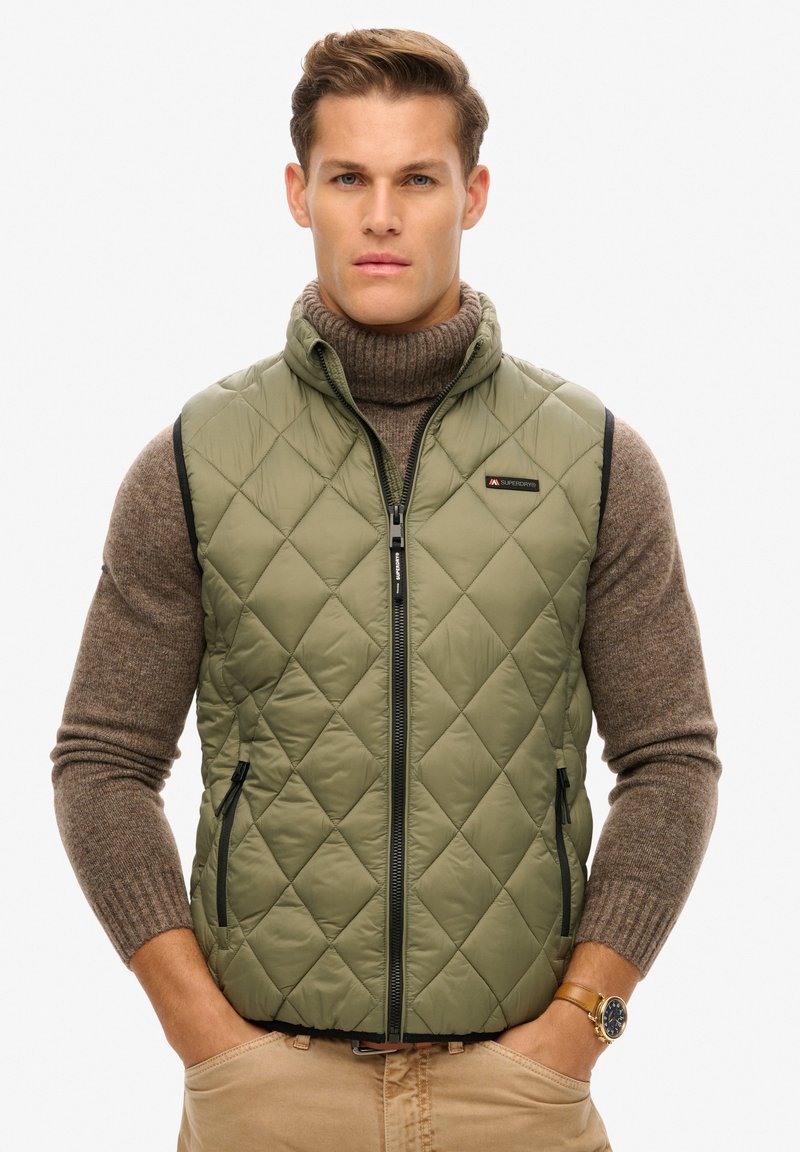 Groene gewatteerde vest met een hoge kraag en ritssluiting, voorzien van zijzakken en zwarte pipings, gedragen over een bruine coltrui.