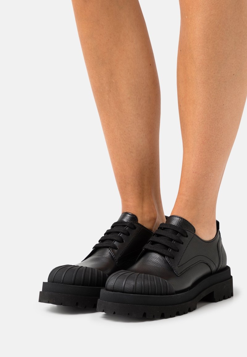 Jambes portant des chaussures montantes en cuir noir avec des embouts en caoutchouc texturés et arrondis, ainsi que des semelles épaisses à crampons, sur un fond clair.