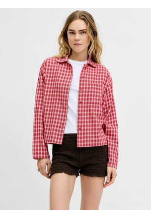 Młoda kobieta w białym t-shircie, czerwonej koszuli w kratę z guzikami i czarnych teksturowanych szortach, stojąca na tle jasnego, jednolitego tła.
