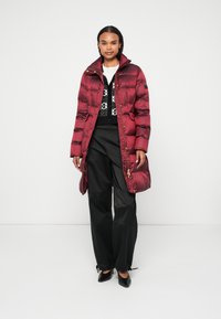 Burgundská péřová bunda s vysokým límcem, ozdobená zlatými detaily, horizontálním švy a bočními kapsami, nošena přes černý vzorovaný svetr.