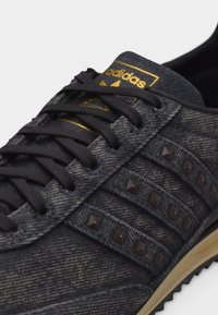 Μαύρο αθλητικό παπούτσι Adidas με μαύρα κορδόνια, τρεις υφασμάτινες ρίγες διακοσμημένες με μαύρα καρφιά και χρυσό λογότυπο Adidas στη γλώσσα.