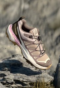 Lys rosa og taupe trail løbesko med tekstureret mesh-overdel, forstærket tå og et distinkt snøresystem. Har en robust ydersål.