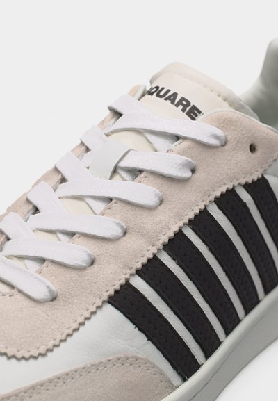 Gros plan sur une chaussure de sport en daim beige et cuir blanc avec des rayures noires et des lacets blancs, nom de la marque partiellement visible sur la languette.
