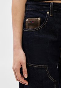 Κοντινό πλάνο ενός χεριού που ακουμπάει πάνω σε σκούρο μπλε τζιν με αντίθεση στα ράμματα και μια μικρή καφέ θήκη με κόκκινο, άσπρο και μπλε λογότυπο στην τσέπη.