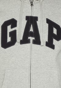 Sudadera gris con cremallera hecha de una tela suave, que presenta un gran logo negro de "GAP" en la parte frontal, con capucha ajustable y bolsillos frontales.