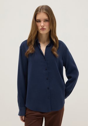 Junge Frau mit langen hellbraunen Haaren, die ein dunkelblaues Hemd zum Knöpfen und eine braune Hose trägt, steht vor einem einfarbigen hellen Hintergrund.