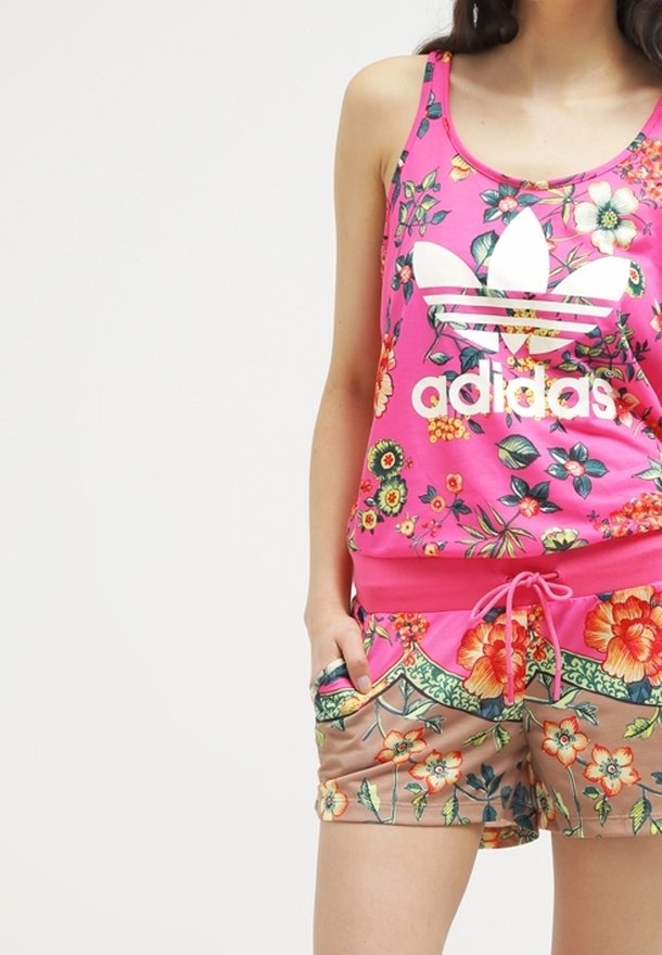 Kvinna i rosa Adidas linne och blommiga shorts med händerna i fickorna mot en enfärgad bakgrund.