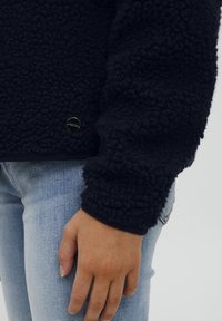 Marineblå fleecejakke med en teksturert, fluffy overflate. Har en rund sølvlogo aksent nær sømmen og ribbestrikkede mansjetter. Kombinert med lyseblå jeans.