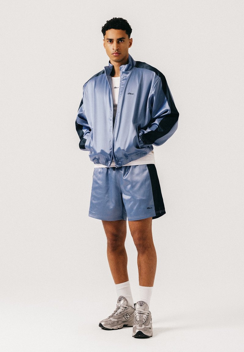 Jeune homme portant un survêtement en satin bleu clair et noir avec des chaussettes blanches et des baskets New Balance grises, debout les mains dans les poches.