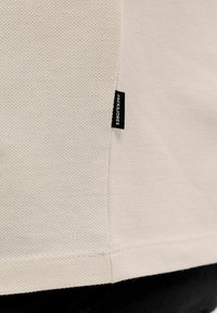 Tissu beige texturé avec une petite étiquette noire indiquant "JACK & JONES" cousue dans une couture verticale près du bord.