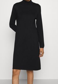 Robe noire à manches longues jusqu'aux genoux avec un col montant, portée par une personne avec un bras plié derrière le dos.