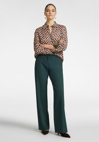 Pantaloni lunghi verdi abbinati a una camicetta a fantasia con forme rosse, blu e beige; la modella è in piedi con le braccia incrociate su uno sfondo neutro.