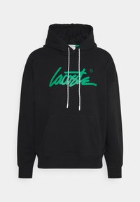 Sudadera negra con capucha, bolsillo frontal y cordones blancos. Presenta un gran logo verde de "Lacoste" en cursiva en la parte delantera.