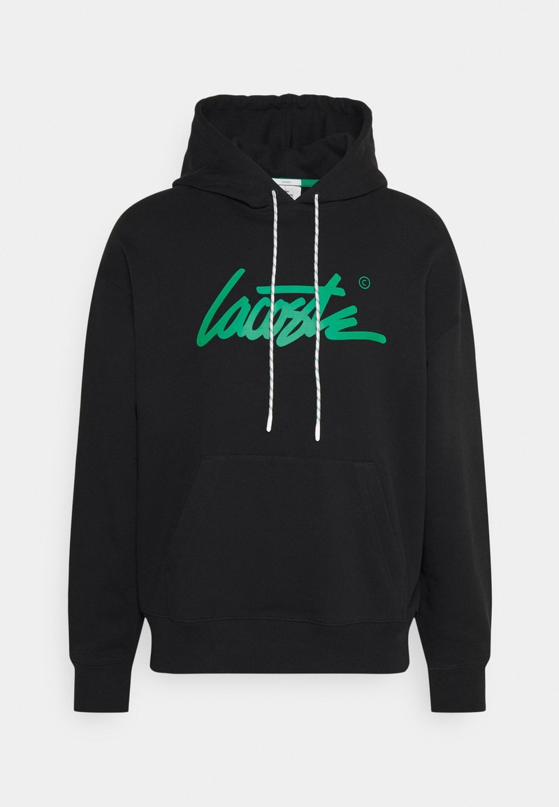 Sudadera negra con capucha, bolsillo frontal y cordones blancos. Presenta un gran logo verde de "Lacoste" en cursiva en la parte delantera.