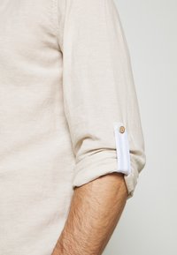 Camisa de lino beige con mangas enrolladas, con un acento de rayas azul y blanca y un detalle de botón de madera. Textura suave, diseño minimalista.