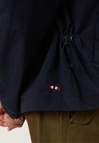 Marinblå jacka med justerbar svart dragsko och liten röd, vit och blå broderad logotyp ovanför fickan på olivgröna byxor.