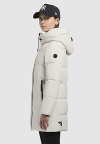 Parka imbottita bianca con cappuccio, dotata di una tasca con zip, patch del logo nero sulla manica e dettagli neri sui lati.