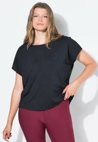 Mladá žena s dlouhými blond vlasy má na sobě volné černé tričko a přiléhavé vínové legíny, pózující proti bílé zdi.