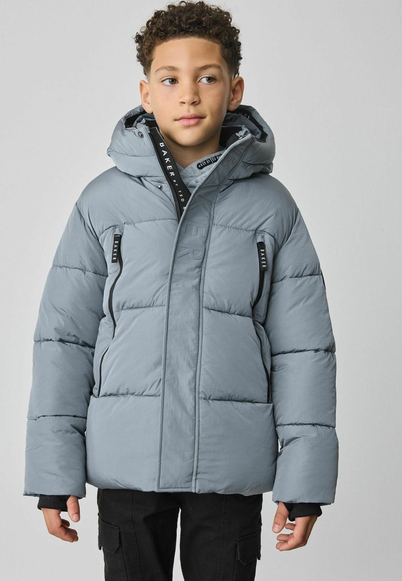Veste matelassée en gris clair, avec une capuche, deux poches zippées à l'avant et des poignets élastiques. Texturée avec des détails matelassés et des étiquettes de marque.