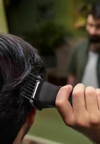 Black hair clipper held in hand, featuring a textured grip and metal blades. Dark hair being styled, with a blurred background.