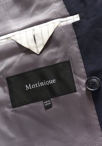 Crni etiketa s natpisom "Matinique" u bijelom tekstu, sivi obrub, dodatna džep u uzorkastoj tkanini i tamna tipka kao detalj.