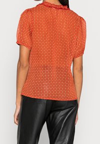 Blusa rossa a fantasia con maniche corte a palloncino e scollatura arricciata, realizzata in tessuto trasparente e con una silhouette aderente. Abbinata a pantaloni in pelle nera.