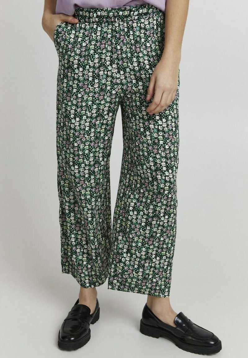 Personne portant un pantalon ample vert à motifs floraux, un haut lavande et des mocassins noirs, debout avec une main dans la poche sur un fond uni.