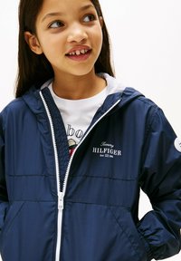 Veste en nylon bleu marine avec une fermeture à glissière blanche, dotée d'une capuche, de poignets élastiques et d'un logo sur la poitrine. Portée par-dessus un T-shirt graphique blanc.