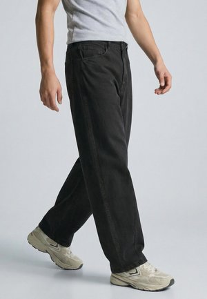 Schwarze Jeans aus Denim mit lockerem Schnitt, geraden Beinen und traditionellen Taschen, kombiniert mit hellen Sneakers und einem grauen T-Shirt.