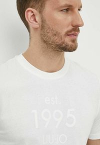 T-shirt bianca con un elegante design stampato che riporta "est. 1995" e "LIU JO" in grigio chiaro. Collo a giro, tessuto in cotone. Vestibilità aderente.