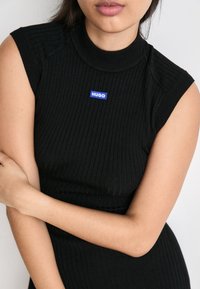 Top sans manches noir côtelé avec un col montant et une étiquette rectangulaire bleue « HUGO » sur le devant. Texture lisse et coupe ajustée.