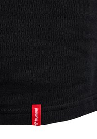 Czarna bawełniana tkanina o teksturowej powierzchni z czerwoną metką logo Hummel w pobliżu dolnego brzegu, prezentująca prosty design i kształt.
