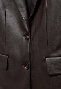 Dunkelbraune Lederjacke mit zwei Schildpattknöpfen, die befestigt sind, und Klappentaschen auf jeder Seite unter der Taille.