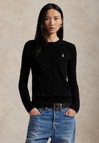 Kvinne med langt svart hår har på seg en svart kabelstrikket genser og blå jeans, med hendene i lommene, mot en ensfarget beige bakgrunn.