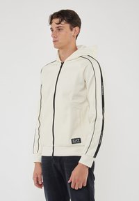 Felpa bianca con cappuccio e zip, dotata di strisce laterali nere e patch con logo, caratterizzata da un design elegante, tessuto leggermente strutturato e tasche frontali.