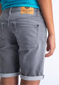 Šedé džínové kraťasy s ohrnutými manžetami, obsahující zadní kapsy a hnědou koženou etiketu na pase. Hladká textura s jemnými švy.