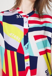 Camicia button-up colorata con motivi geometrici in rosso, blu, aqua, giallo e grigio. Include numeri visibili e un design con colletto a fascia.