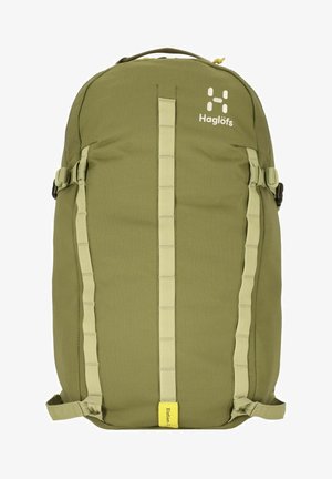 Mochila verde oliva con una superficie de tela suave, correas en la parte frontal y una etiqueta destacada amarilla en la parte inferior. El logo de la marca es visible.