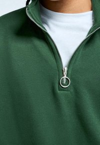 Felpa verde con zip e colletto alto, dettagli in argento. Il tessuto sembra avere una texture morbida e viene indossata sopra una maglietta bianca.