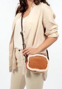 Borsa a tracolla in suede marrone con bordo in pelliccia bianca, dotata di una lunga tracolla regolabile e chiusura con zip, tenuta da una persona.