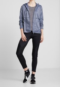 Veste légère à capuche avec fermeture éclair en bleu ardoise portée sur un haut gris, associée à des leggings noirs moulants et des chaussures de sport noires.