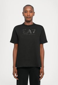 Cilvēks ar īsiem matiem valkā melnu EA7 Emporio Armani t-kreklu un melnas bikses, stāvot pret vienkāršu gaišu fonu.
