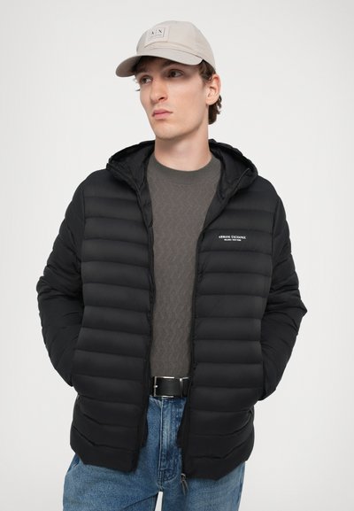 Veste matelassée noire avec un quilting horizontal, capuche et fermeture éclair ; portée sur un pull gris texturé, associée à un pantalon en denim bleu et une casquette beige.