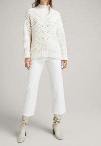 Nainen, joka on pukeutunut kermaiseen palmikkoneuleiseen neuleeseen, valkoisiin leveisiin nilkkapantoihin ja beigeihin löysiin korkokenkiin, seisoo vaaleanharmaalla taustalla.