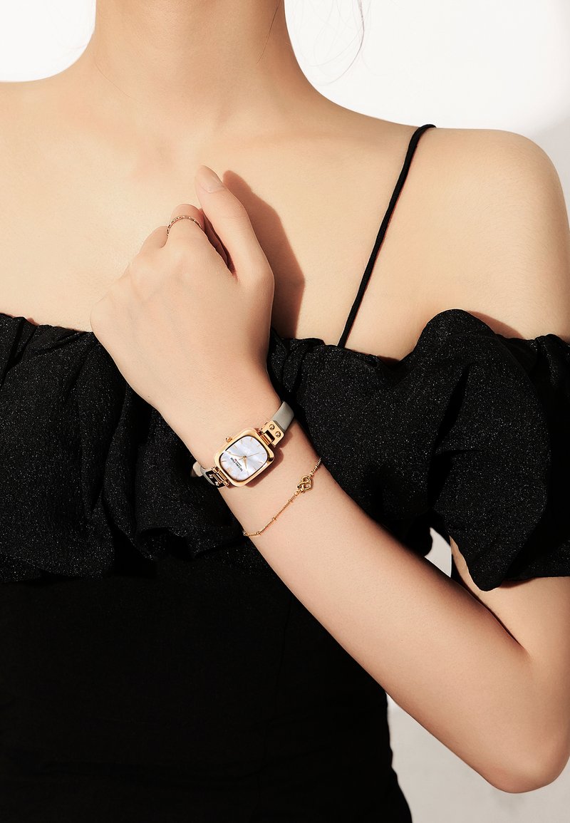 Montre rectangulaire avec un cadran blanc et des accents dorés, bracelet noir et gris clair. Un délicat bracelet en or avec un petit pendentif en forme de cœur au poignet.