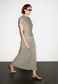 Gestreiftes, locker sitzendes Kleid in Beige und Schwarz, mit kurzen Ärmeln und einem gestuften Saum. Das Modell trägt Sonnenbrillen und schwarze Flip-Flops.