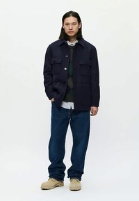 Jaunas vyras stovi su tamsiai mėlynu švarku, sluoksniuota megztiniu ir marškiniais, laisvomis mėlynomis džinsomis ir smėlio spalvos batais prieš baltą foną.